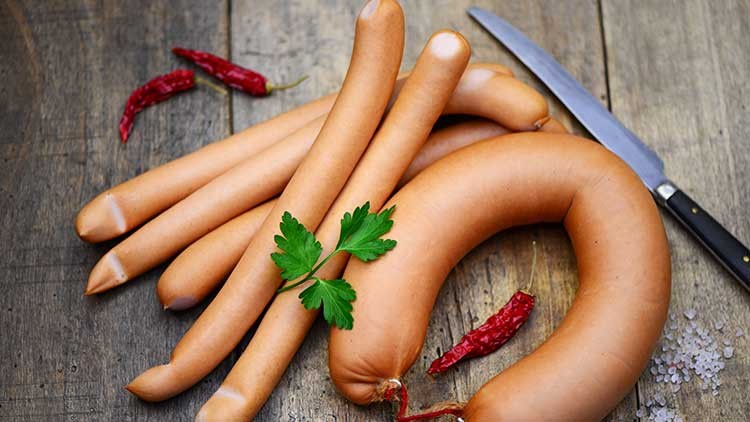 Mehrere Würstchen und eine Ringwurst liegen auf Holz, dekoriert mit Petersilie, roten Chilischoten und Salz, daneben ein Messer.