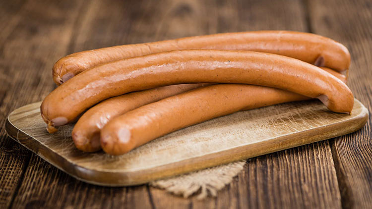 Vier braune, gekochte Würste liegen übereinander auf einem hölzernen Schneidebrett mit mattem Holzuntergrund.