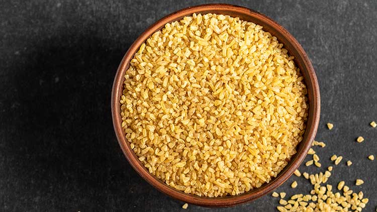 Holzschale gefüllt mit gelbem Bulgur auf dunklem Untergrund, einige Körner liegen verstreut daneben.
