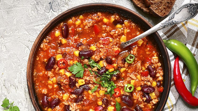 Eine Schüssel Chili con Carne mit Kidneybohnen, Mais, Hackfleisch, Paprika, frischen Kräutern und Chilischoten, daneben Brot.