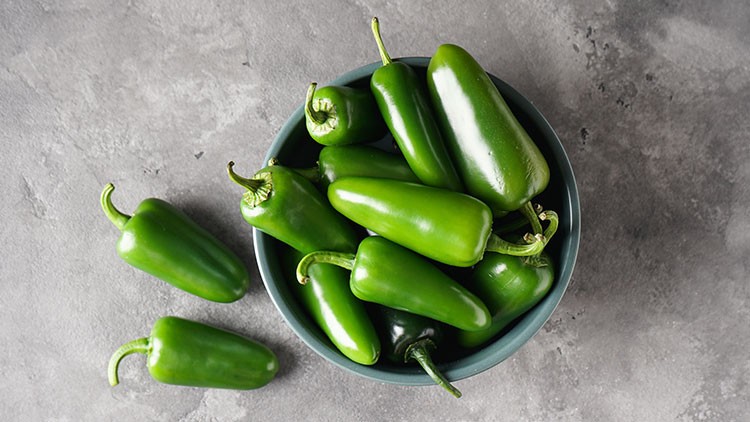 Grüne Jalapeños liegen teilweise in einer dunklen Schüssel und teilweise auf einer grauen Steinoberfläche.