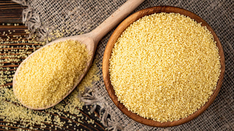 Eine Holzschale und ein Holzlöffel mit gelbem Couscous liegen auf einem Untergrund aus grobem Leinenstoff.