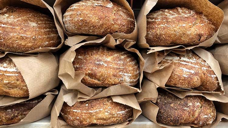 Mehrere goldbraune Brote liegen übereinander in braunen Papiertüten arrangiert.