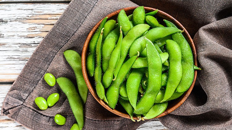 Grüne Edamame-Bohnen liegen in einer Holzschale auf einem braunen Tuch, einige Bohnen sind daneben auf dem Tisch verteilt.