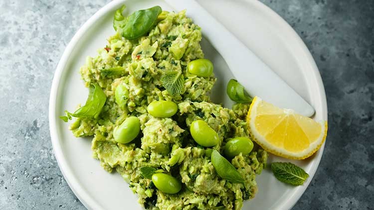 Guacamole mit Edamame-Bohnen, frischen Kräutern und Zitronenscheibe auf einem weißen Teller mit weißem Messer serviert.
