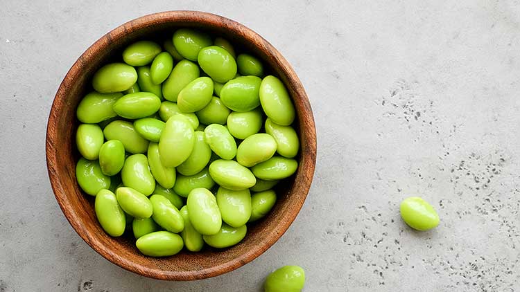 Eine hölzerne Schüssel ist mit leuchtend grünen Edamame-Bohnen gefüllt, eine Bohne liegt daneben auf grauem Untergrund.