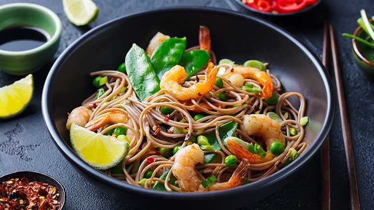 Schwarze Schale mit gebratenen Garnelen, Vollkornnudeln, Erbsen, Zuckerschoten, Limettenspalten und roten Chiliflocken.