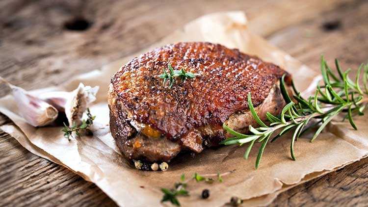 Ein gebratenes Stück Fleisch mit knuspriger Kruste liegt auf Backpapier, dekoriert mit frischen Kräutern, Knoblauch und Rosmarin.