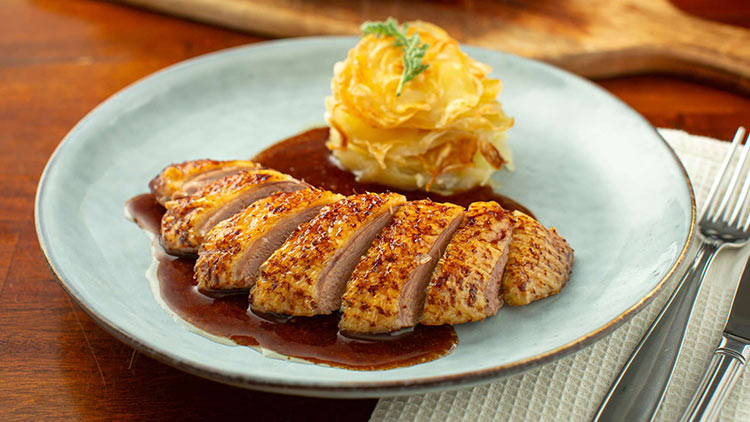 Aufgeschnittene gebratene Entenbrust in dunkler Sauce, serviert mit Kartoffelgratin auf einem hellblauen Teller.