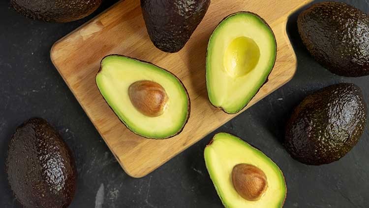 Auf einem Holzbrett liegen halbierte Avocados mit Kern sowie ganze Avocados auf einer dunklen Unterlage.