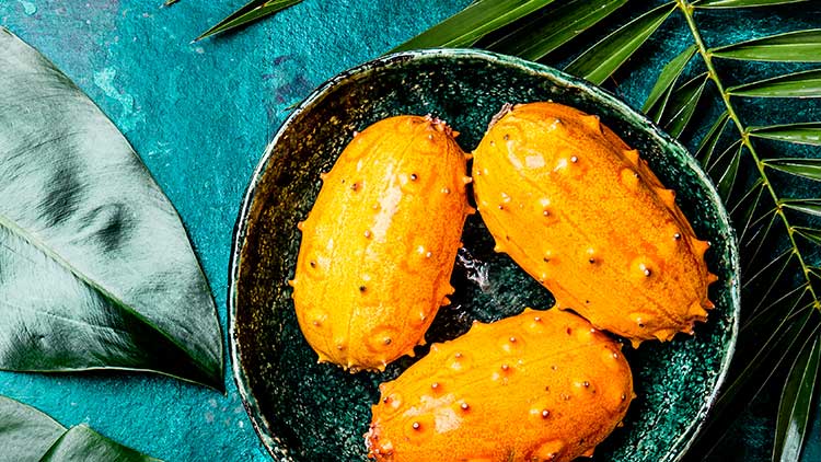 Drei orange Kiwano-Früchte mit stacheliger Schale liegen in einer dunkelgrünen Schale auf türkisfarbenem Untergrund mit Blättern.