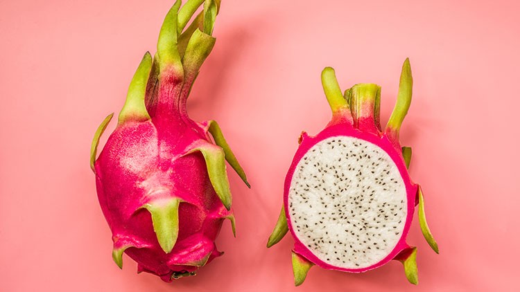 Eine ganze und eine aufgeschnittene pinke Drachenfrucht mit weißem Fruchtfleisch und schwarzen Samen liegen auf rosa Hintergrund.