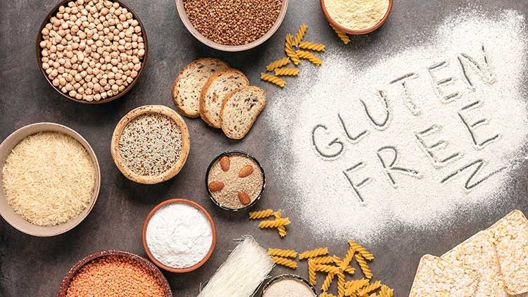 Verschiedene glutenfreie Lebensmittel und Schrift „GLUTEN FREE“ aus Mehl auf einer dunklen Fläche, darunter Nudeln und Brot.