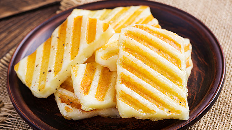 Goldbraune Halloumi-Streifen mit Grillmuster liegen auf einem dunklen Teller aus Holz.
