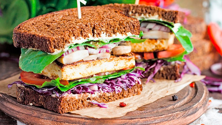 Ein Sandwich mit dunklem Brot, Salat, Tomaten, Rotkohl, Zwiebeln und gegrilltem Käse liegt auf Holzuntergrund mit Papier.