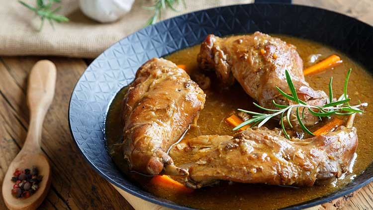 Drei gegarte Geflügelkeulen in brauner Sauce mit Karottenstiften und Rosmarin in einer schwarzen Schale, daneben ein Holzlöffel.