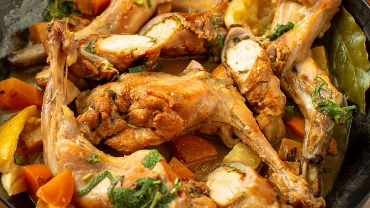 Geschmorte Kaninchenkeulen mit Karotten, Kartoffeln und Kräutern in einer schwarzen Pfanne, daneben ein Schälchen Kartoffelpüree.