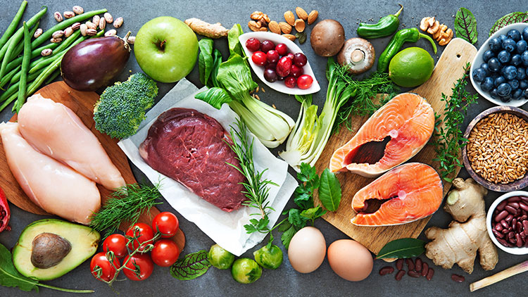 Verschiedene frische Lebensmittel wie Gemüse, Obst, Hülsenfrüchte, Fleisch, Fisch und Gewürze auf einem Tisch arrangiert.