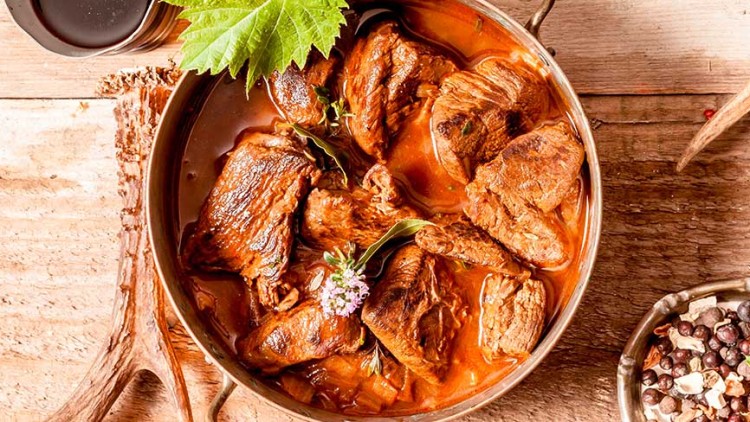Kupferfarbener Topf mit geschmortem Fleisch in Soße, dekoriert mit Kräutern, daneben Gewürze, Tannenzweig und ein Hirschgeweih.