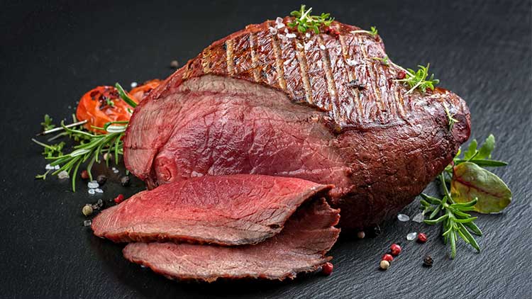 Ein Stück gebratenes Fleisch mit Kräutern, Pfefferkörnern, grobem Salz und einer Scheibe Tomate auf schwarzem Schiefer.