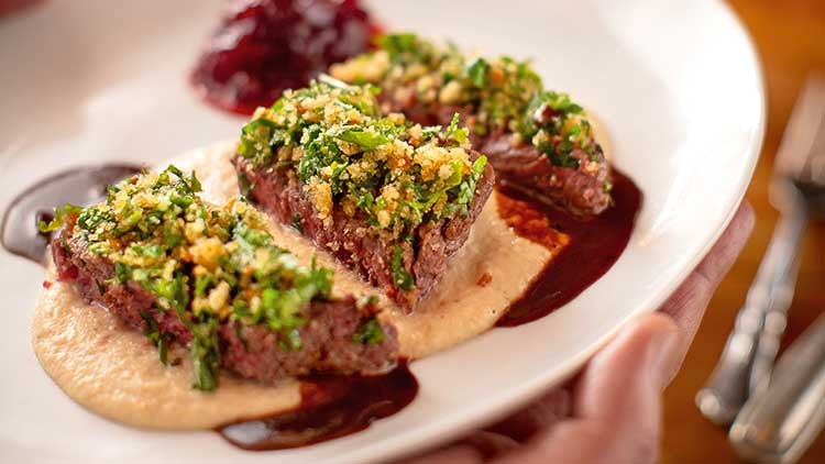 Zwei Scheiben Fleisch mit grüner Kruste auf cremigem Püree, dunkler Soße und rotem Fruchtkompott auf weißem Teller.