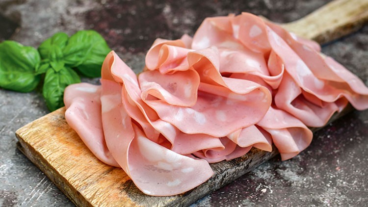 Mehrere Scheiben Mortadella liegen auf einem Holzbrett, daneben frische grüne Basilikumblätter.