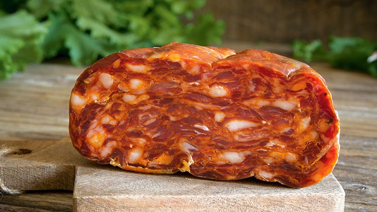 Aufgeschnittene rote Wurst mit sichtbarer Marmorierung liegt auf einem Holzbrett, im Hintergrund grüne Blätter.