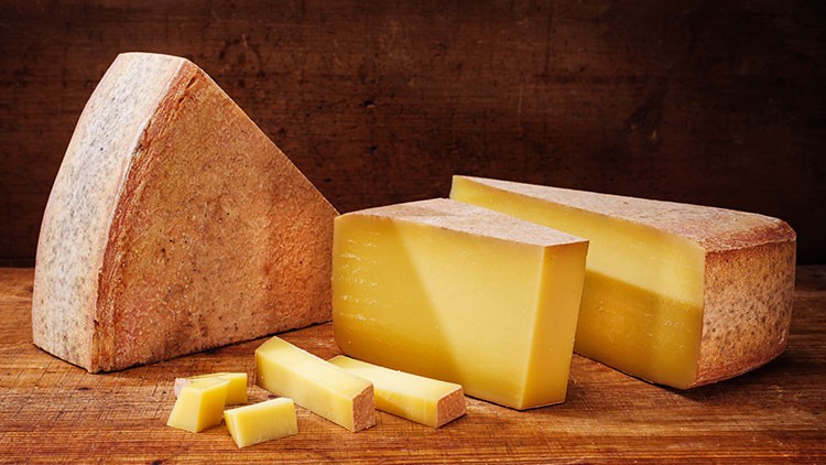 Mehrere Stücke Hartkäse in verschiedenen Größen liegen auf einer Holzunterlage vor einem braunen Hintergrund.
