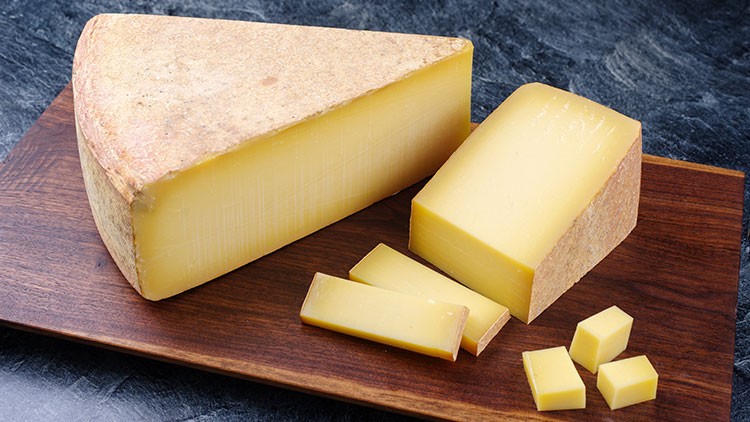 Ein großes und ein kleineres Stück Hartkäse mit Rinde sowie Käsestücke und Würfel liegen auf einem Holzbrett.
