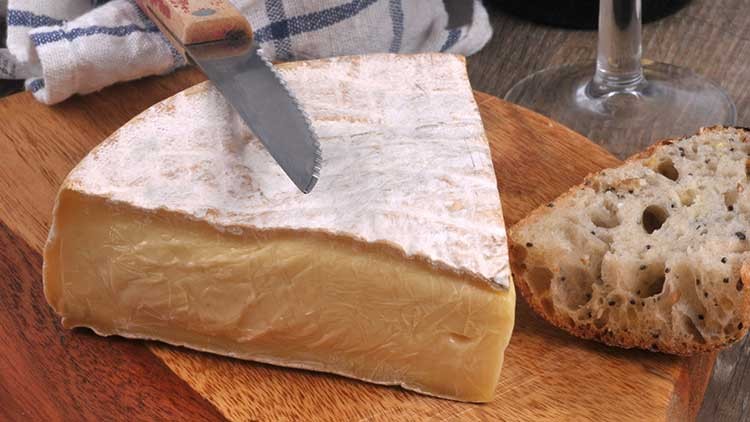 Ein Stück Käse mit weißer Rinde liegt auf einem Holzbrett, daneben ein Brotscheibchen und ein Messer mit Holzgriff.
