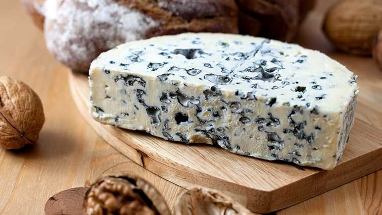 Ein Stück Blauschimmelkäse liegt auf einem Holzbrett, umgeben von Walnüssen und dunklem Brot.