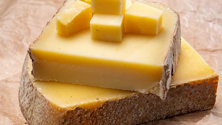Ein Stück Hartkäse mit Naturrinde liegt auf Packpapier, darauf liegen mehrere kleine Käsewürfel.