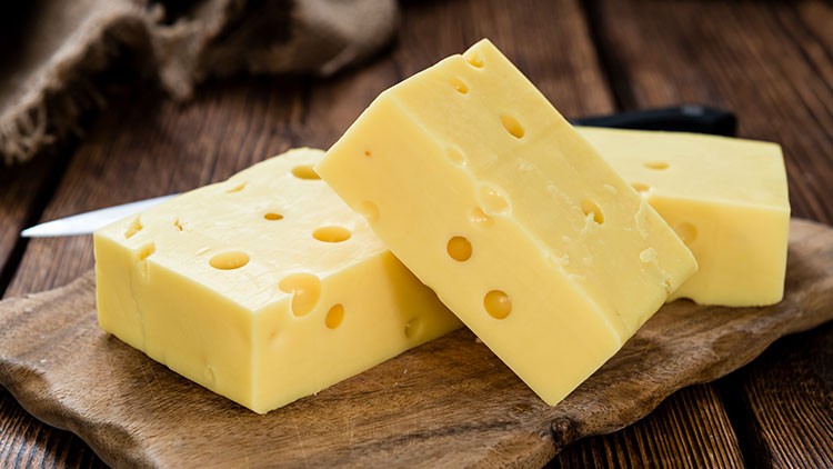 Zwei Stücke gelber Käse mit Löchern liegen auf einem hölzernen Schneidebrett, im Hintergrund ein Messer und Stoff.