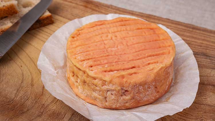 Ein runder Weichkäse mit orangefarbener Rinde liegt auf Backpapier auf einem Holzbrett, daneben liegt ein Messer mit Brot.