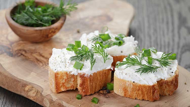 Drei Brotscheiben mit Frischkäse, frischem Dill und geschnittenem Schnittlauch auf einem Holzbrett, daneben eine Schale Dill.