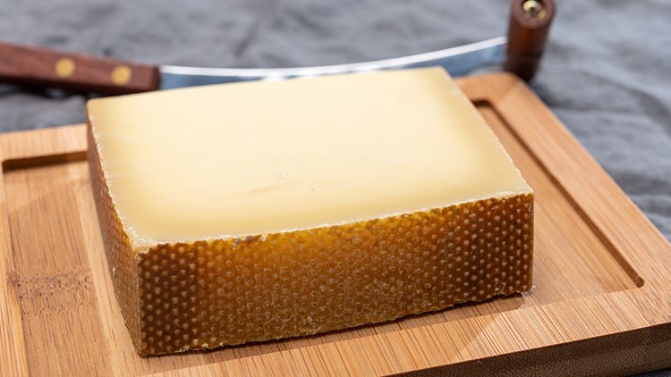 Ein rechteckiges Stück Käse mit brauner Rinde liegt auf einem Holzbrett, im Hintergrund ein Käsemesser.