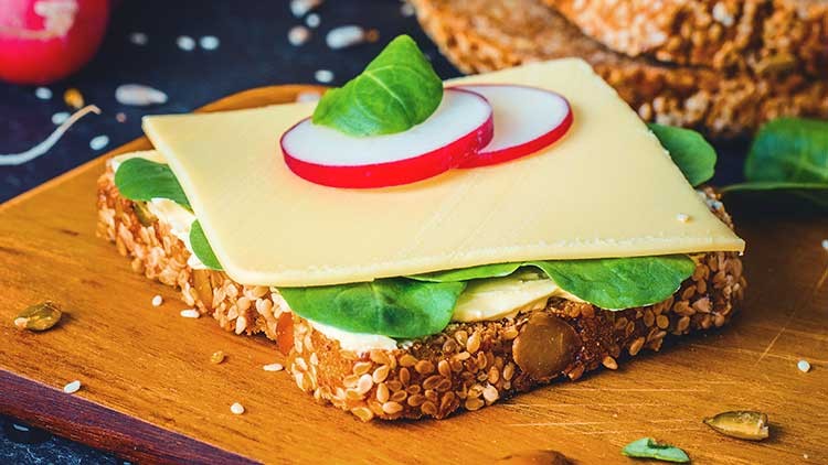 Ein belegtes Körnerbrot mit Käse, Blättern frischen Spinats, Radieschenscheiben und Kürbiskernen auf einem Holzbrett.