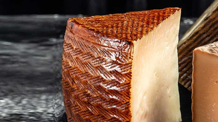 Ein keilförmiges Stück Hartkäse mit brauner Rindenstruktur und hellem Inneren liegt auf einer schwarzen Oberfläche.