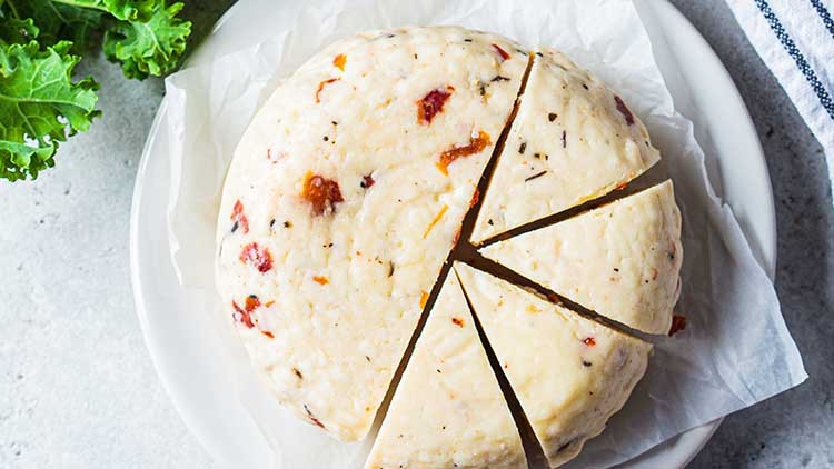 Runder Käse mit Kräutern und roten Stücken, auf weißem Teller und Backpapier, teilweise in Stücke geschnitten, daneben grüne Blätter.