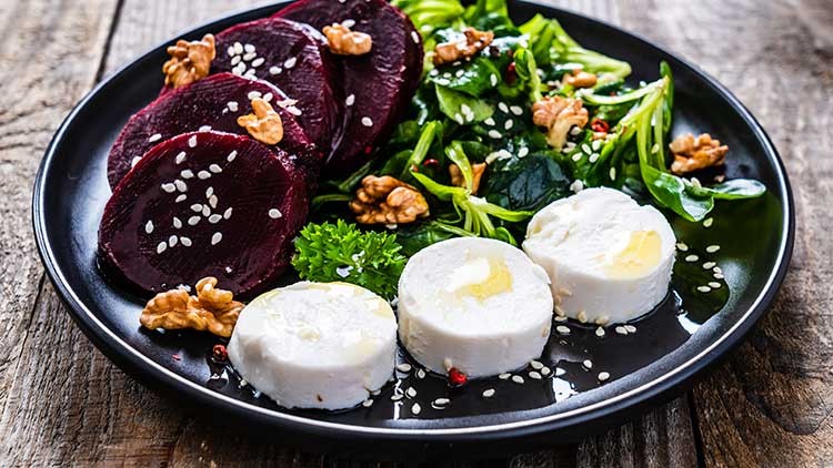 Schwarzer Teller mit Salat, Walnüssen, Rote-Bete-Scheiben, Sesam und drei Stücken Ziegenkäse mit Öl beträufelt.