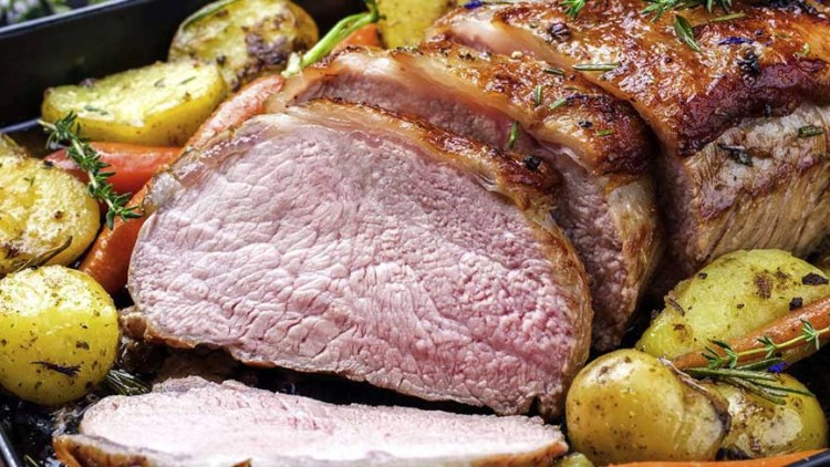 Ein aufgeschnittener Braten liegt in einer Backform, umgeben von Kartoffeln, Karotten, Zwiebeln und frischen Kräutern.