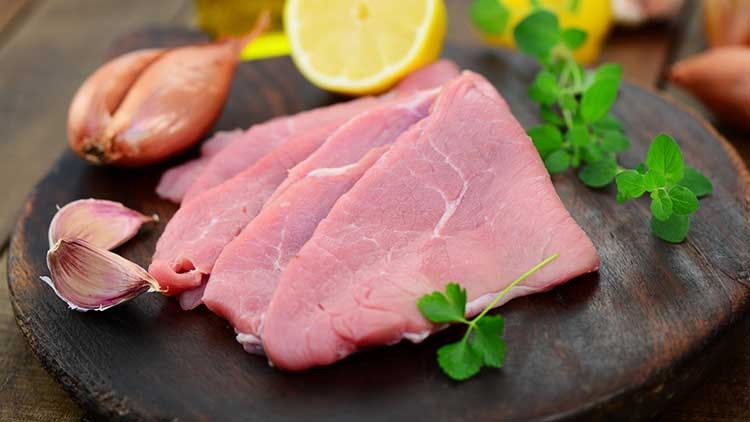 Dünne, rohe Fleischscheiben auf dunklem Holzbrett mit Knoblauchzehen, Schalotten, Kräutern und einer halbierten Zitrone.