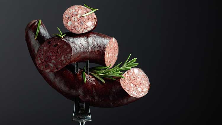 Eine aufgeschnittene dunkle Wurst mit Rosmarinzweigen auf einer Gabel vor dunklem Hintergrund.
