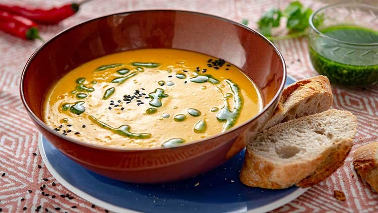 Orange Suppe in brauner Schale mit grünem Öl und schwarzen Samen, daneben Brotscheiben auf blauem Teller und Glas mit grünem Dip.