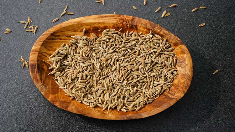 Kleine braune Kümmelsamen liegen in einer ovalen Holzschale auf schwarzem Untergrund, einige Samen sind außen verstreut.