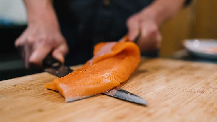Ein Lachsfilet wird auf einem Holzbrett mit einem Messer zerteilt, Hände halten das Filet und das Messer fest.