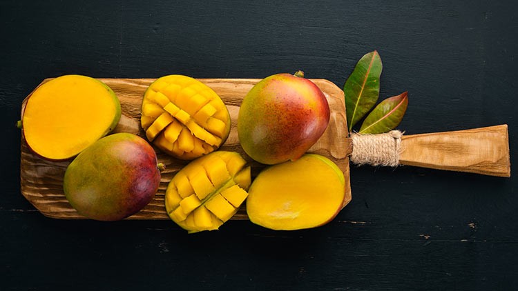 Ganze und aufgeschnittene Mangos liegen auf einem Holzbrett mit Griff, dekoriert mit grünen und roten Blättern, vor dunklem Hintergrund.