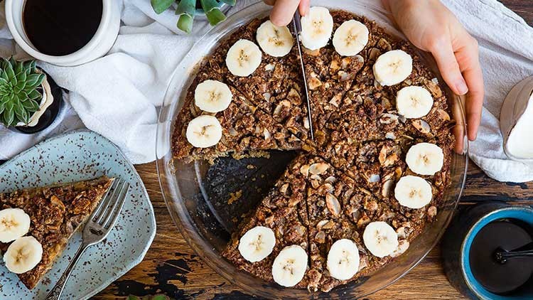 Ein rundes Bananen-Nuss-Dessert in einer Glasform wird angeschnitten, umgeben von Kaffee, Tellern und grüner Dekoration.