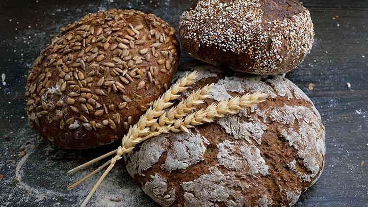 Drei Brotlaibe mit Samen- und Körnertoppings sowie zwei Ähren liegen auf dunklem Untergrund.