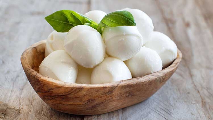 Kleine Mozzarellakugeln liegen in einer Holzschale, dekoriert mit frischen Basilikumblättern, auf einer hellen Holzoberfläche.